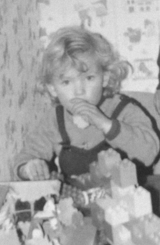 Schwarzweißfoto von blond gelocktem Kind in Latzhose beim Lego bzw Duplo Bauen vor 70er-Kinderzimmer-Tapete