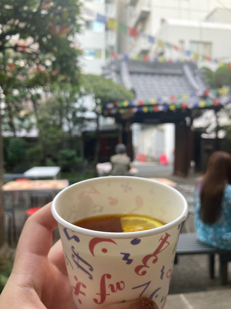 手前にはカップを持つ手　中にはホットレモネード　サフランで色濃いオレンジ色　向こうには寺の境内の様子　ベンチや机が並べられ　門までのあいだに木と木の間にカラフルなちいさな旗がたくさんかかっている