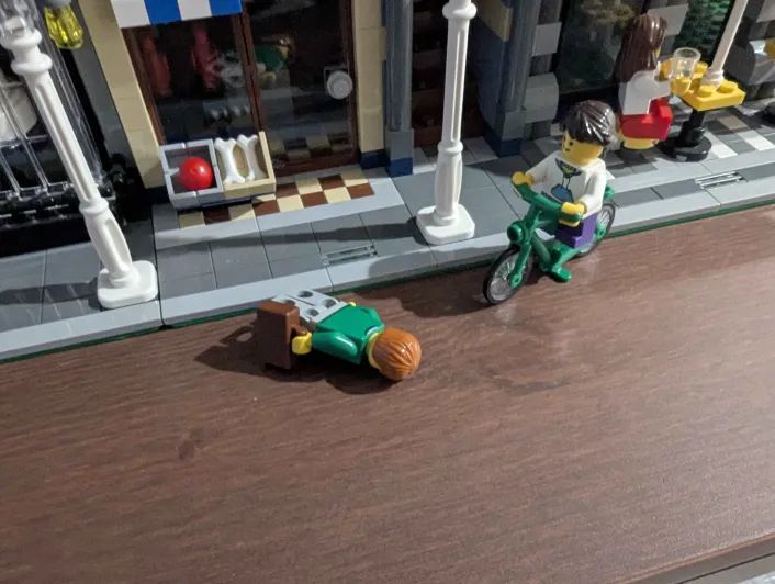 A LEGO minifigure wearing a green sweater and grey pants and holding a brown briefcase lies face-down after falling off the sidewalk.