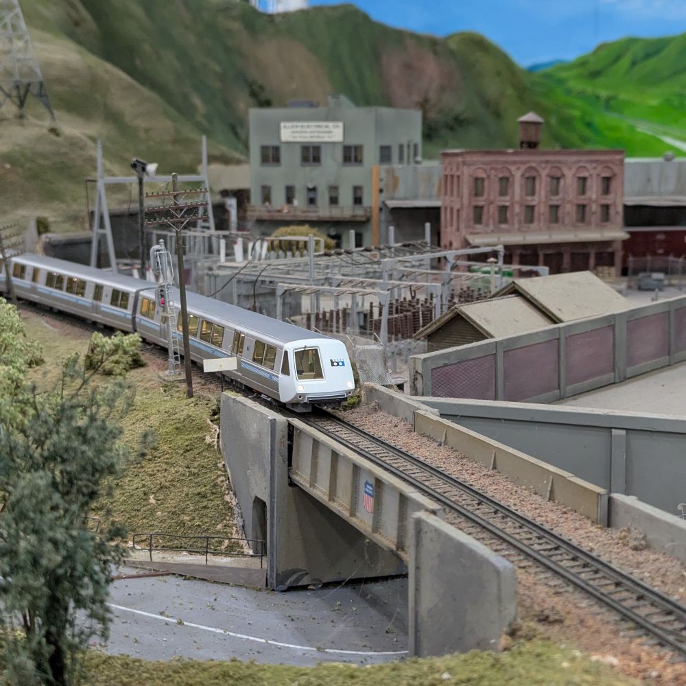 An HO scale BART train - a sleek silver and white metro train with a pointed nose - crosses a concrete and steel bridge on a model train layout. An electrical substation and brick factories are behind it, with hills and a blue sky in the background.