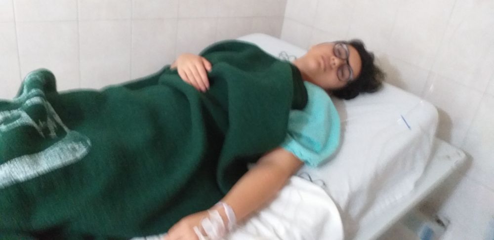 Blurry picture of a person asleep on a hospital bed, thick blanket covering their body except their arms, an IV connected one arm