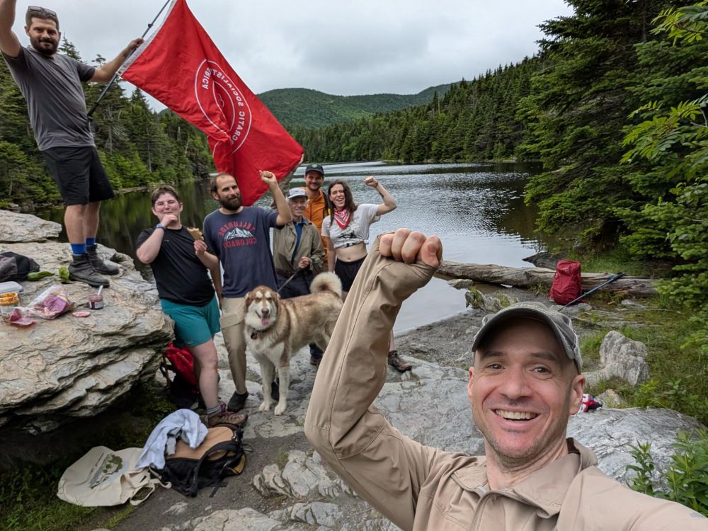 A group of socialists raising fists by a lake with a DSA flag and a very wolfy happy dog