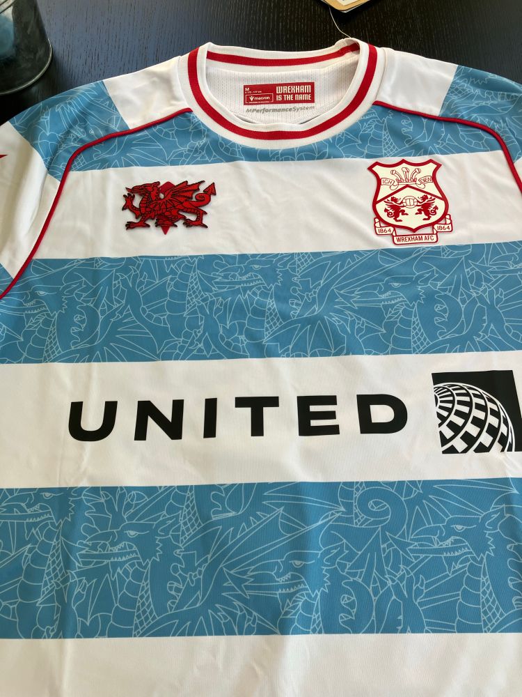 The front of the Wrexham AFC third kit. It has light blue stripes with outlines of dragons on a white background, the United Airlines logo in black, and near the top, the Wrexham badge in red and the red dragon from the Welsh flag