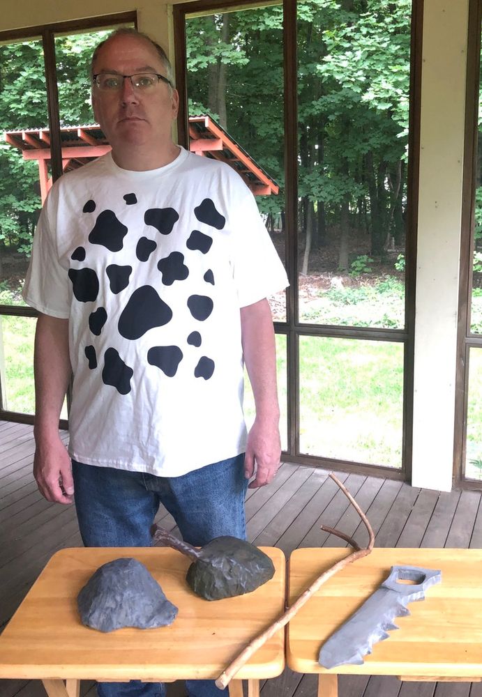 A man in a spotted cow pattern shirt stands in front of a table. On the table are four primitive looking tools. It is a real-life tribute to the Gary Larsen Far Side "Cow Tools" cartoon. 