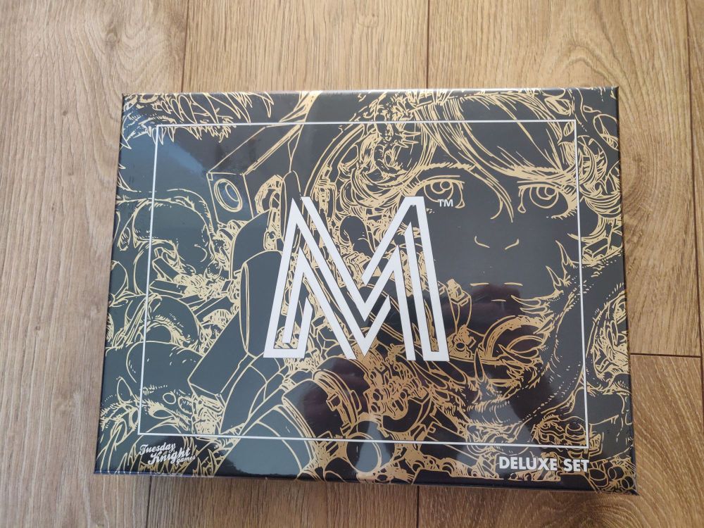 The Mothership RPG deluxe box set. A shiny black box with gold linework art and a large white M logo.