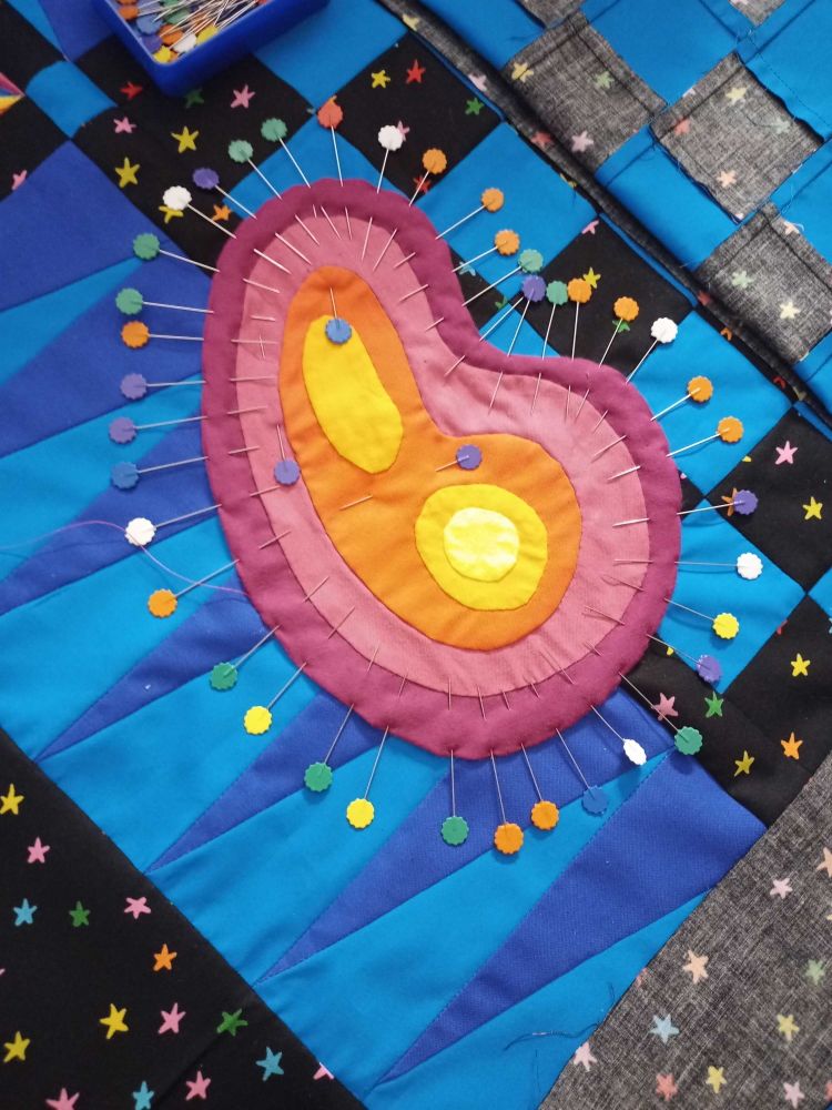 close up photo of a quilt. an organic blob shape of layered coloured fabric has been meticulously pinned with like a billion pins to the quilt top.