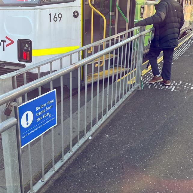 A tram platform stop with a notice saying "No low-floor trams at this stop. Call PTV on 1800 800 007 if you need assistance". A person with a walking stick is preparing to board a high floor tram.