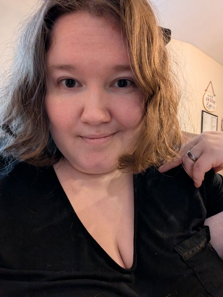 Kit, a person with messy gold-brown hair and ample cleavage, looks at the camera with a crooked smile. If you look closely, you can see cat hair on their black clothing. 