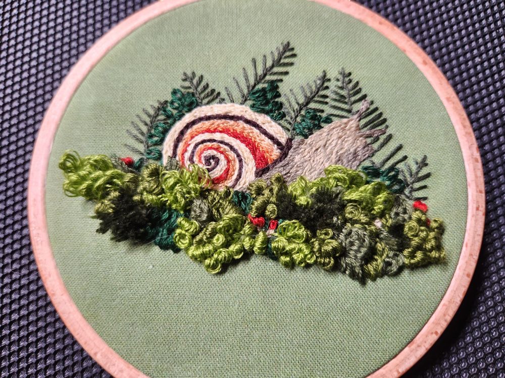 An embroidered realistic snail with a background of ferns and a foreground of dimensional stitched mosses. The piece is about 4 inches diameter and embroidered on green cotton cloth. 