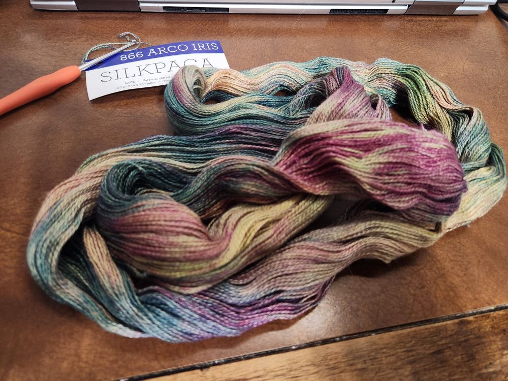A loosened skein of hand-died multicolor yarn is laying on a brown wooden desk.  The yarn is 70%baby alpaca and 30%silk, and the muted rainbow colors have a beautiful sheen.