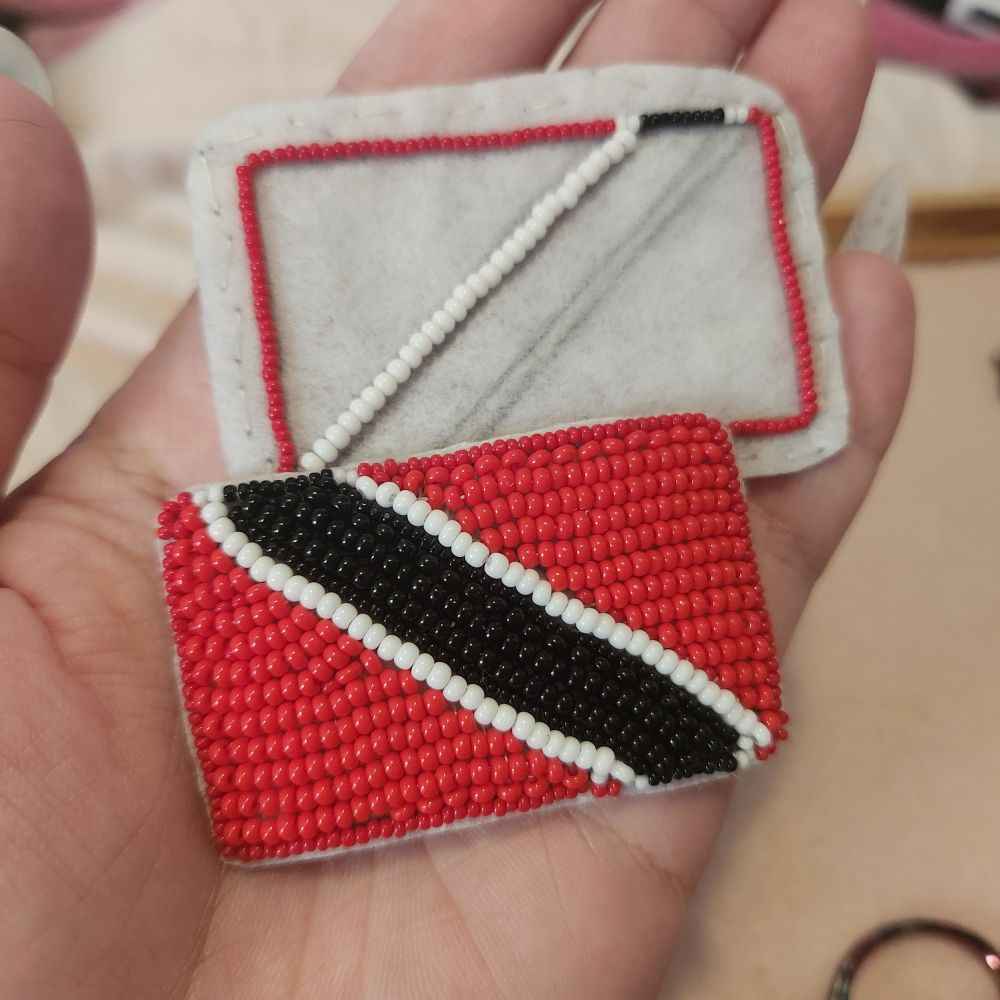 Flat beaded version of the Trinidad and Tobago flag, it is a red flag with a black stripe diagonally across it with two small white stripes on either side of the black. 