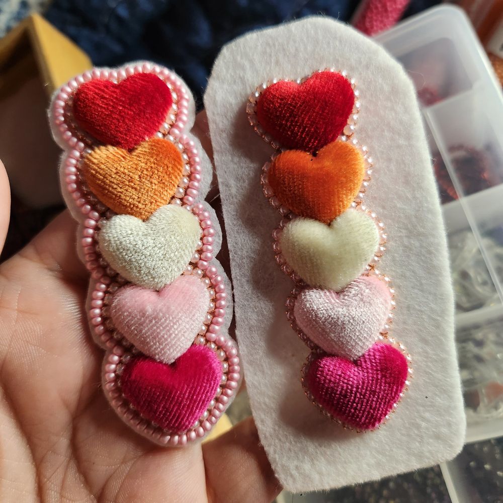 Small heart shaped cabachons in a row, making the lesbian flag. Red, orange, white, light pink & fuchsia. There is rhinestone pearl banding around the hearts, they are in the process of being made. 
