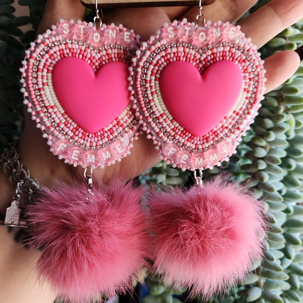 Large beaded earrings, a pink heart cabachon is the middle of the earrings. Lines of beadwork in alternating rows, featuring pink, red & cream beads surround it. There is a line of rhinestone banding as well. Pink alphabet letter beads spell out stone at the top of the earring and femme at the bottom. A pink rabbit fur pom pom is attached at the bottom of the earring. They are hanging on silver earring hooks.