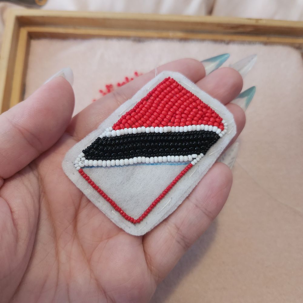 Flat beaded version of the Trinidad and Tobago flag, it is a red flag with a black stripe diagonally across it with two small white stripes on either side of the black. The beadwork is unfinished. 