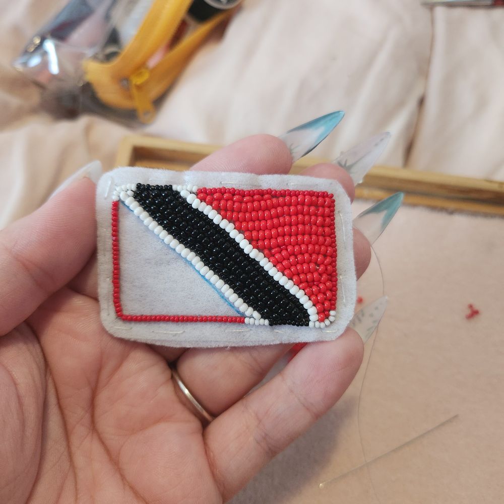 Flat beaded version of the Trinidad and Tobago flag, it is a red flag with a black stripe diagonally across it with two small white stripes on either side of the black. The beadwork is unfinished. 
