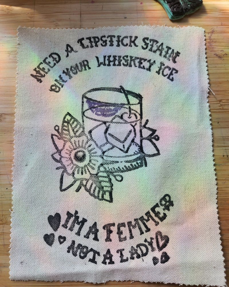 Block print stamp of a whiskey glass with a lipstick print on it, there is ice and a cherry in the whiskey glass, beside the glass is a bold six petaled flower. Above the glass are the words "Need a lipstick stain on your whiskey ice." 
Below the glass are the words "I'm a femme, not a a lady." 
The print is in black ink, the lipstick stain is purple and it is printed on cream canvas cloth. 