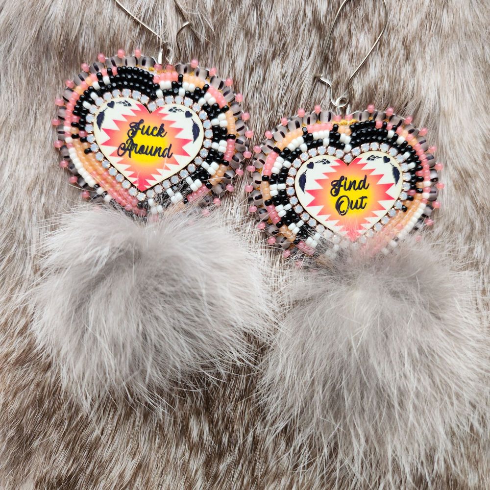 Beaded cabachon earrings one half says "fuck around" and the other half says "find out". They have cream, black, neon orange and neon pink in a striped and miss match pattern. They have mottled brown rabbit fur pom poms attached at the bottom. The background is rabbit fur 
