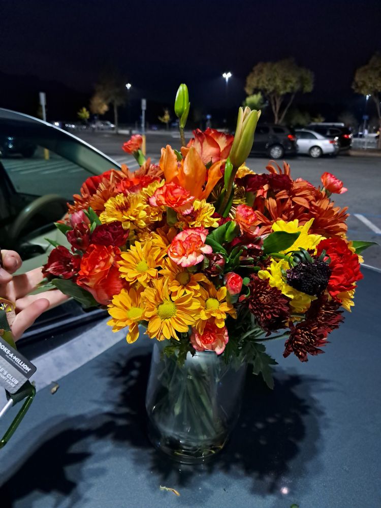 Flower arrangement of red, orange, and yellow, sitting on top of a car hood in a parking lot.