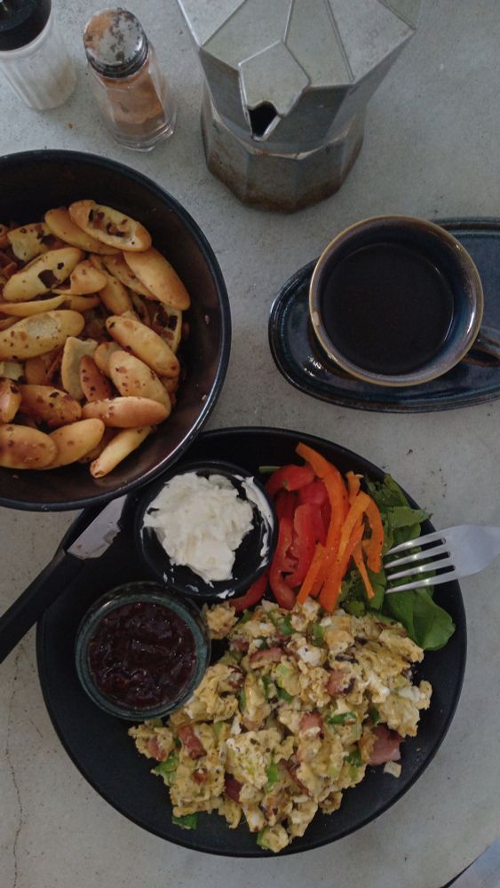Foto de cima de uma mesa de mármore com uma cumbuca com bolachas ovais amanteigadas, um saleiro, um potinho com canela, uma cafeteira moka de alumínio, um pires oval com uma xícara cheia de café e no meio um prato com ovos mexidos com bacon, uma saladinha de tomate, pimentão e rúcula com duas molheiras, uma com geleia e outra com labneh.
