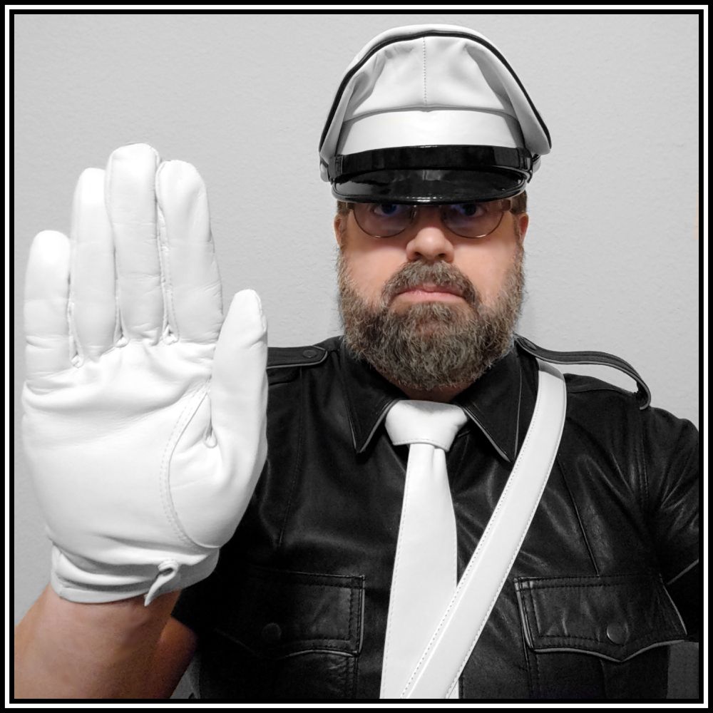 A selfie of Roldán Belenos wearing a black leather shirt, white leather tie, white Sam Browne belt, and a white Muir cap with black trim; he is holding up his right hand in a 'stop' gesture.