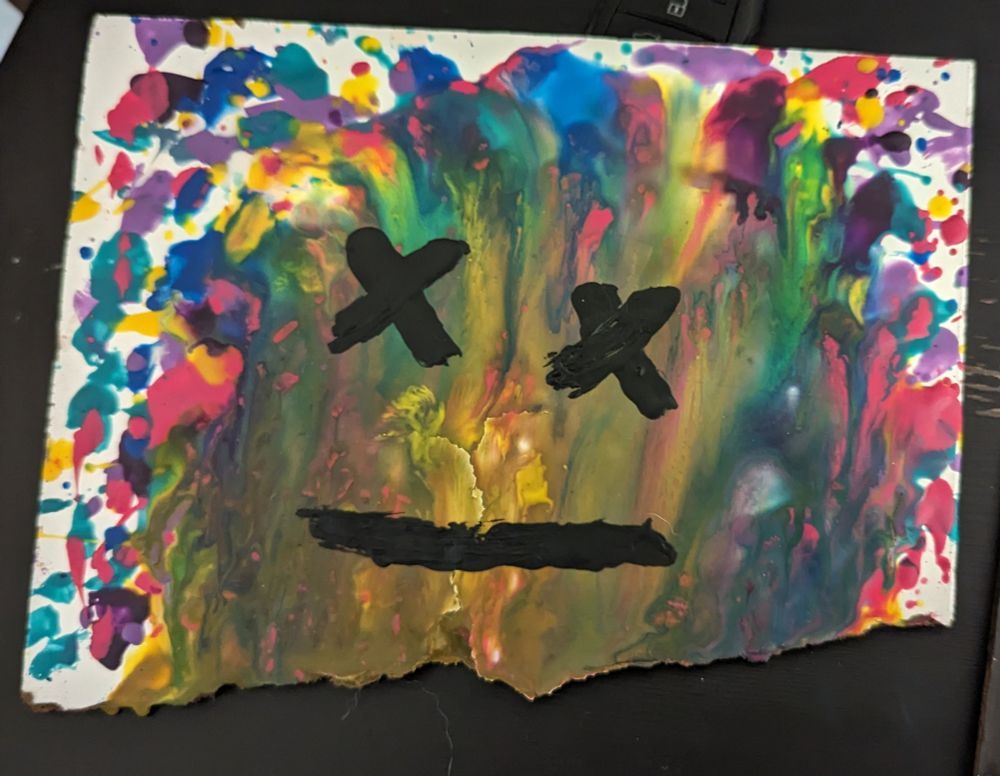 A broken whiteboard slate with a waxy rainbow color burst emanating from a crack. Nearest to the crack the wax is yellow. Two black X marks form eyes with a horizontal black segment forming a mouth to a face atop the color burst