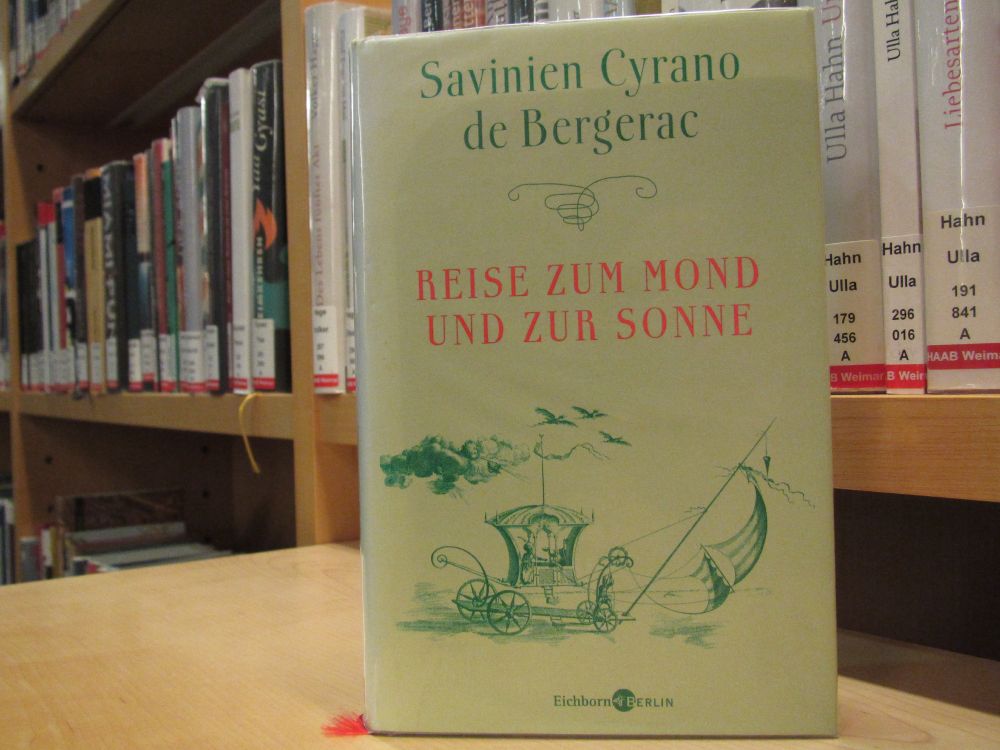 Ein Foto des Buchs "Reise zum Mond und zur Sonne" von Savinien Cyrano de Bergerac. Das Buch hat einen hellen, grünlichen Schutzumschlg, auf dem mittig in roter Schrift der Buchtitel und darüber in grün der Name des Verfassers steht. Auf den unteren Drittel des Covers ist die Illustriation einer historischen Zeichnung einer Kutsche mit Segel abgebildet.
Das Buch steht aufrecht auf einer Ablage aus hellem Holz, im Bildhintergrund Bücherregale der Romanbibliothek der HAAB Weimar.