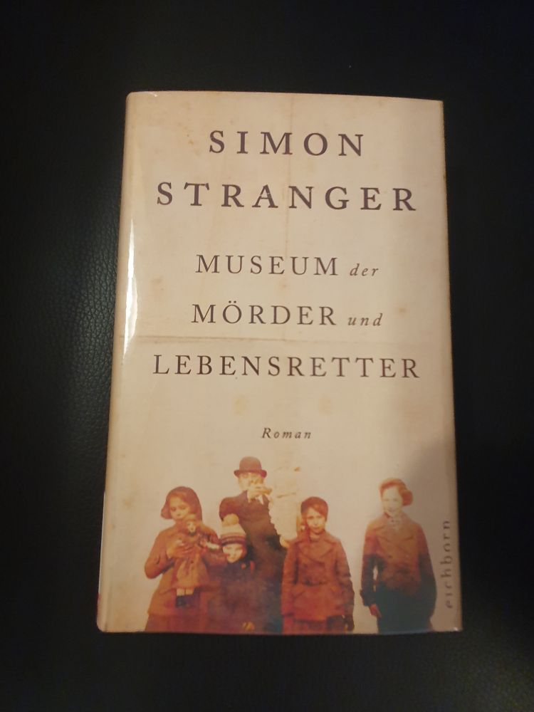Abgebildet ist Simon Strangers Roman "Museum der Mörder und Lebensretter" liegend auf schwarzem Hintergrund. Das Cover zeigt eine Familienfotografie.