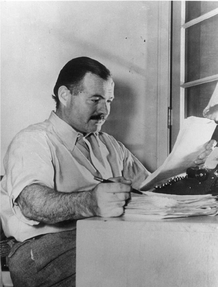 Auf der schwarz-weiß-Fotografie ist Ernest Hemingway im Profil, sitzend an seinem Schreibtisch, zu sehen.