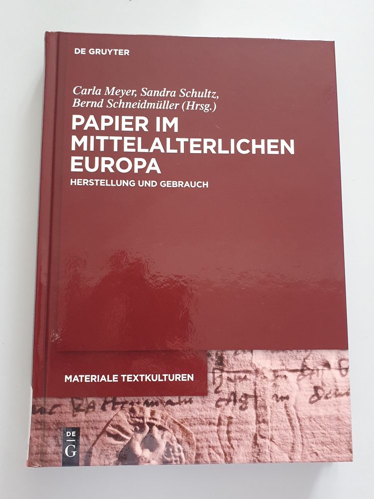 Foto des Buches "Papier im mittelalterlichen Europa : Herstellung und Gebrauch / hrsg. von Carla Meyer, Sandra Schultz und Bernd Schneidmüller". Das Buch hat ein weinrotes Hochglanzcover mit Titel, Verlagsangabe und Herausgeberangabe in weißer Schrift. Am unteren Rand des Einbandes ist das Fragment einer mittelalterlichen HAndschrift abgebidet.