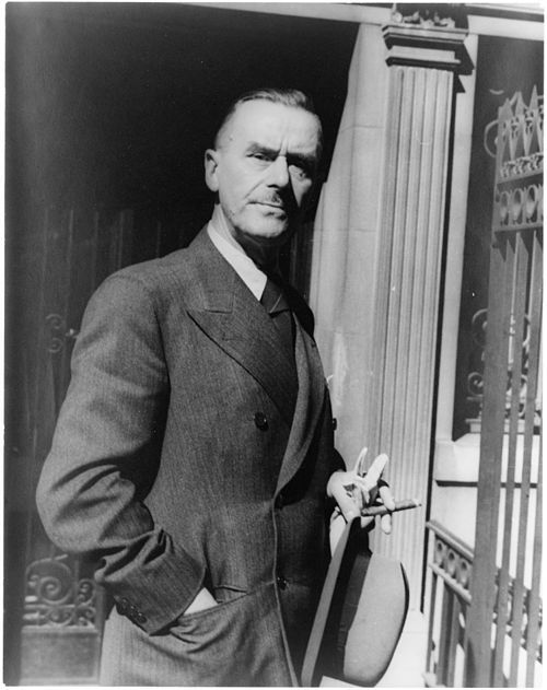 Schwarz/Weiß-Portrait von Thomas Mann. Man sieht nur die obere Hälfte seines Körpers. Er trägt einen Anzug oder einen Mantel; eine Hand steckt in der Tasche der Jacke, in der anderen Hand hält er einen Hut und eine Zigarre.