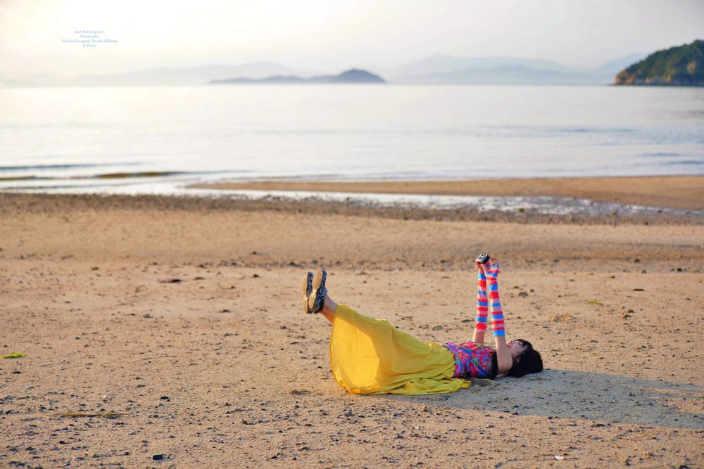 穏やかな海の砂浜でカメラの写ルンですを持って仰向けになって両手両足を上げている女性