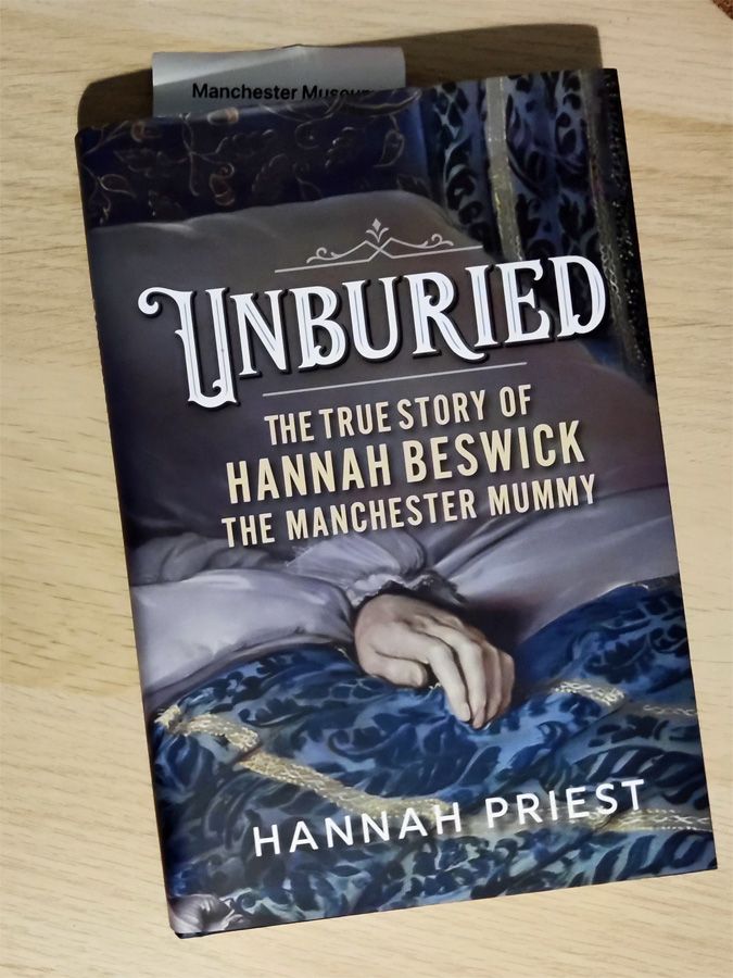 A hardback book on a light pine table. The cover art shows bed coverings with an exposed hand of an woman. 