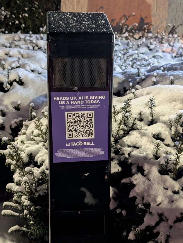 A fast food drive through speaker surrounded by snowy bushes.  There is a purple sign with a QR code on it that says: HEADS UP. AI IS GIVING US A HAND TODAY.