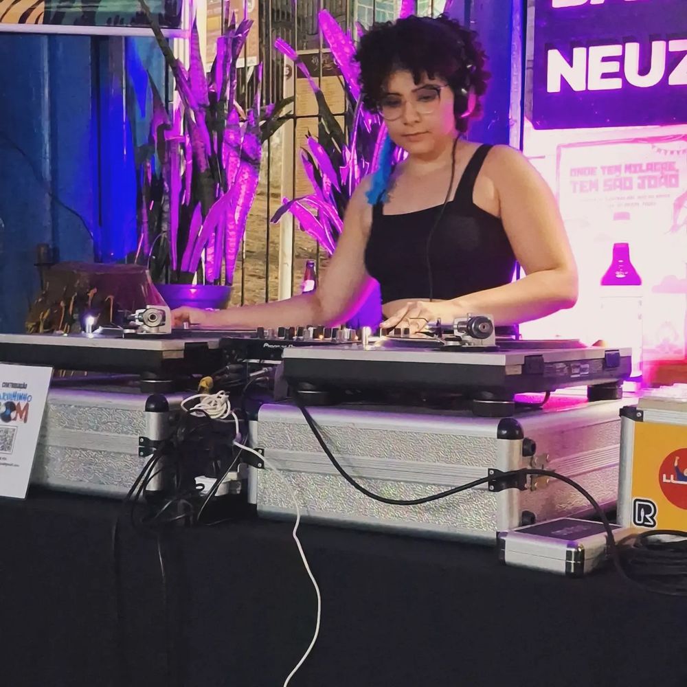 Eu discotecando. Apareço atrás de dois toca-discos e um mixer. Tenho pele clara, uso óculos e tenho cabelo cacheado escuro. Estou usando um top preto.