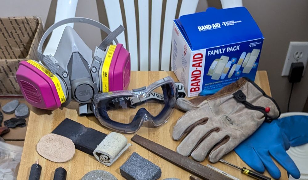 Photo of flintknapping saftey gear. 3M Half Mask Respirator, Goggles, Band-aids, Two Leather Palm Pads, Rubber Palm Pad, Leather Gloves, Nitrile Gloves