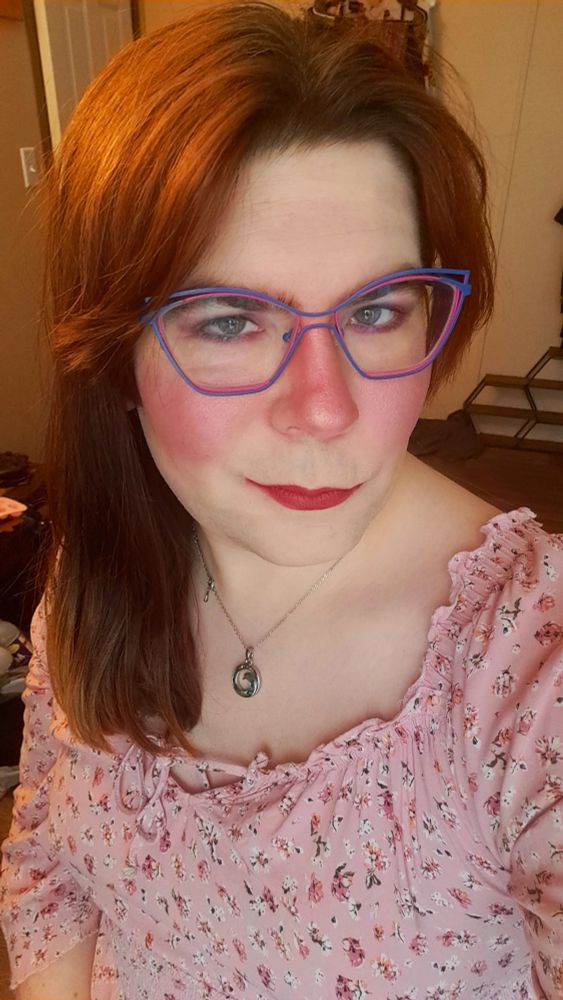 A selfie of the Refuge Psystem. They have redbrown hair and bi flag colored glasses. Their top is pink with a floral pattern. Their cheeks and nose are rosey from the makeup and there's a bit of sparkle to it. They have red lips in a pursed smirk and a necklace with a swirling green stone pendant. Their eyes are green.