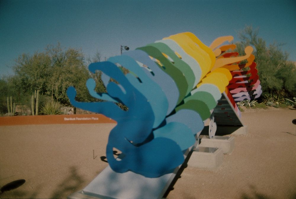 A photo of a line of 15 or so abstract metal figures made from flat metal and in a variety of colors, forming a rainbow. They are outdoors in a desert botanical garden. 