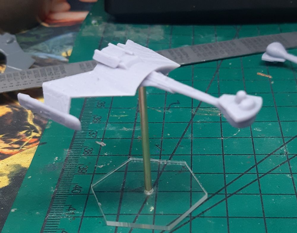 A WIP 3D printed miniature of a Klingon K'tinga class ship. The model is mounted to a clear hexagonal base by a piece of brass rod.