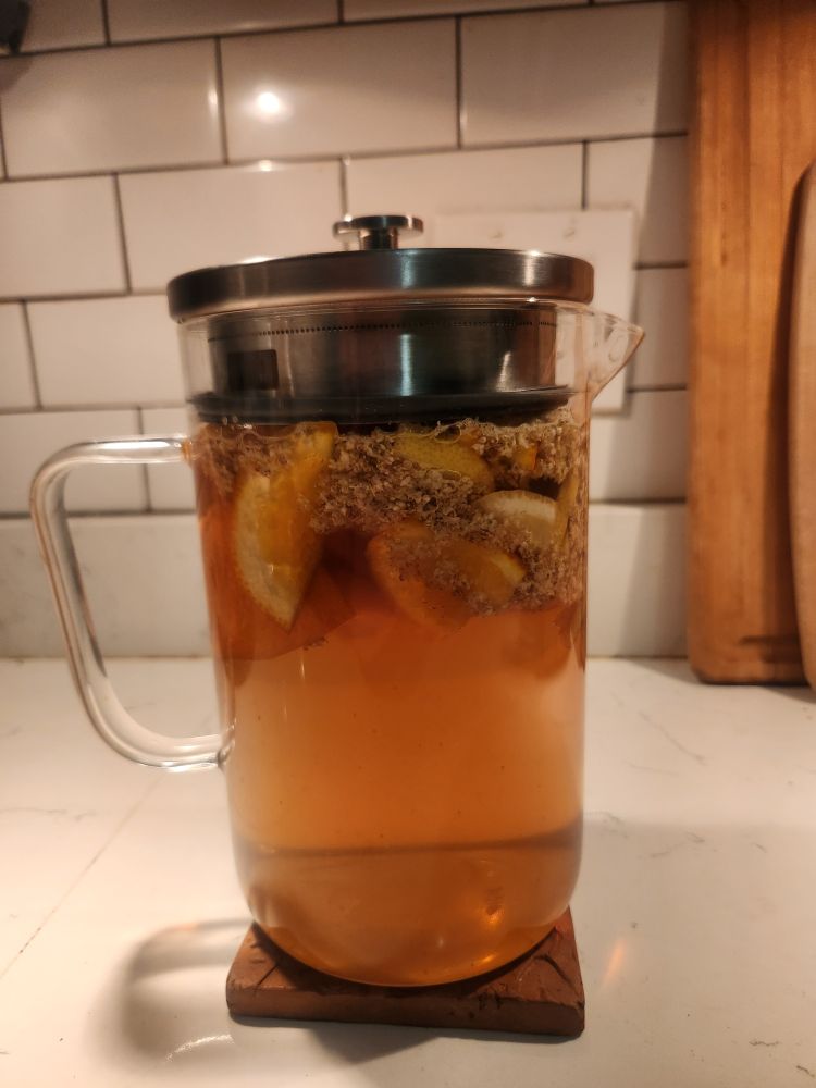 A glass pitcher.  In it,  elderflower blossoms and chunks of lemon and orange float in a copper colored liquid. 