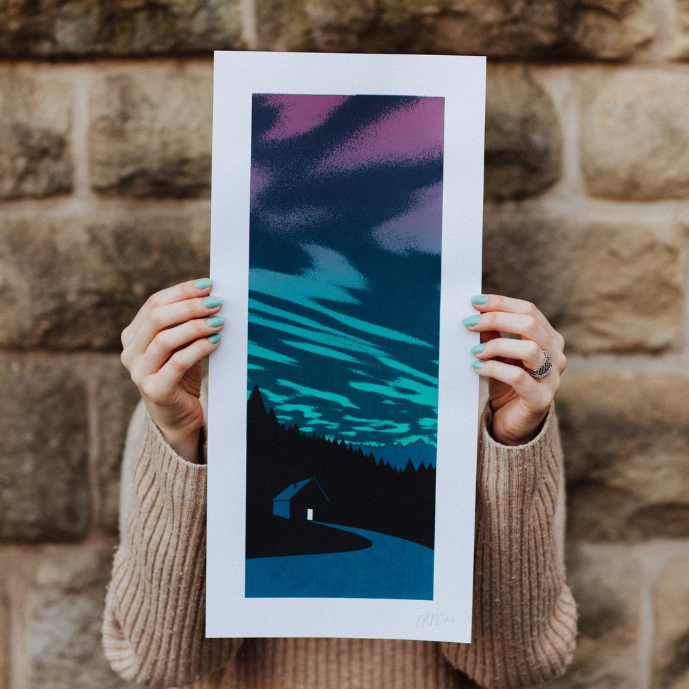 A screenprint of a cabin in the woods with the Northern Lights in the background. The print is being held in two hands.