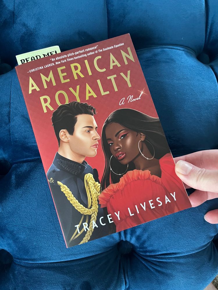 A photo of Tracey Livesay’s romance novel, American Royalty, set on a piece of quilted blue velvet furniture. 