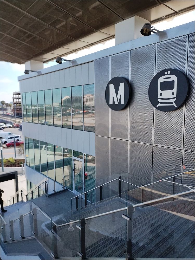 Bike Hub at the LAX station. Image shows a glass & steel mesh building beside a stair set which ascends three levels with a small view of a parking lot on the right and other buildings in the distance.