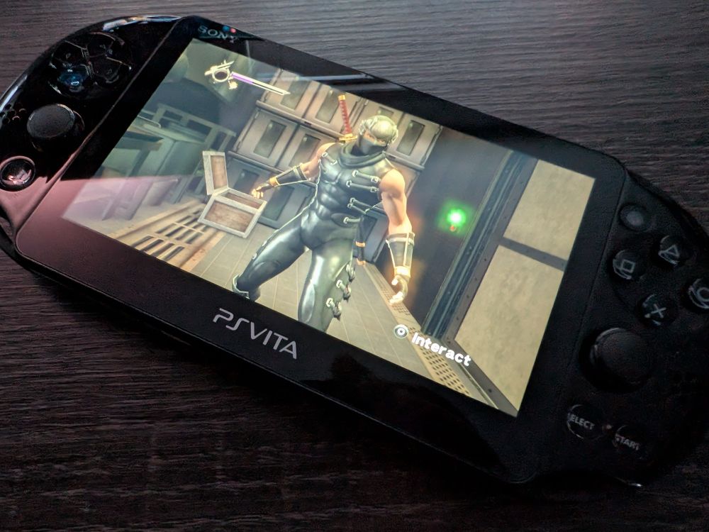 A Playstation Vita sits on a dark wood table. On the screen is Ninja Gaiden.