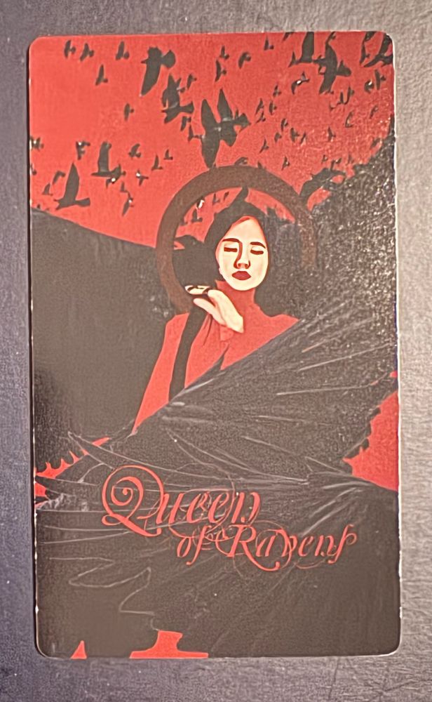 The Queen of Ravens shows a woman wearing red, eyes closed, on a black and red background made up of small and large ravens. Card by 7DA. 