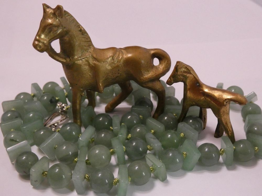the green aventurine necklace coiled in a circle around two small brass figurines of a mare and a foal. necklace has a pattern of alternating round and square beads. 