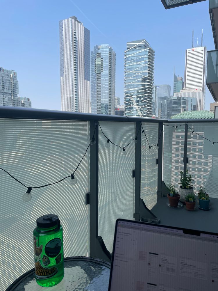 View from the deck chair of a balcony railing covered in string lights. A water bottle and laptop screen corner are in the foreground with a city skyline in the background.