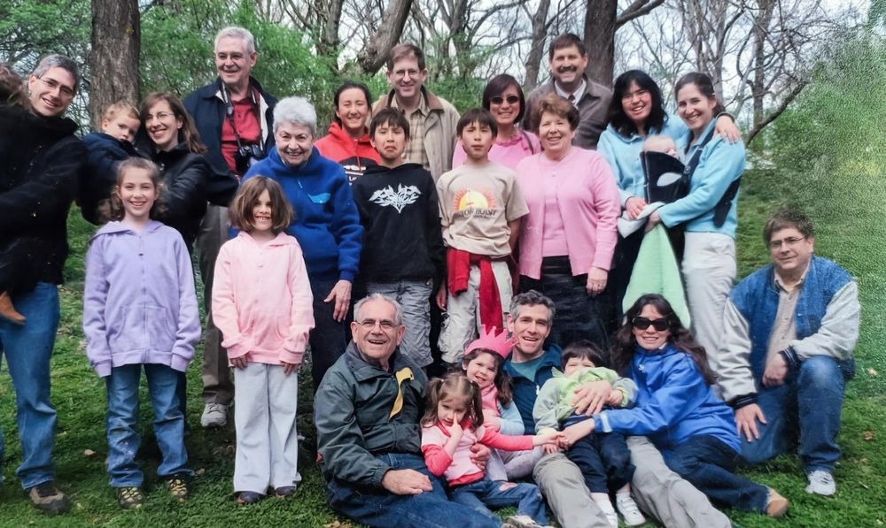 Photo of many white people smiling at the photographer. The people range from young to old. Some are sitting on the ground, many are standing. There is greenery in the background. The photo looks to have been taken in a park.