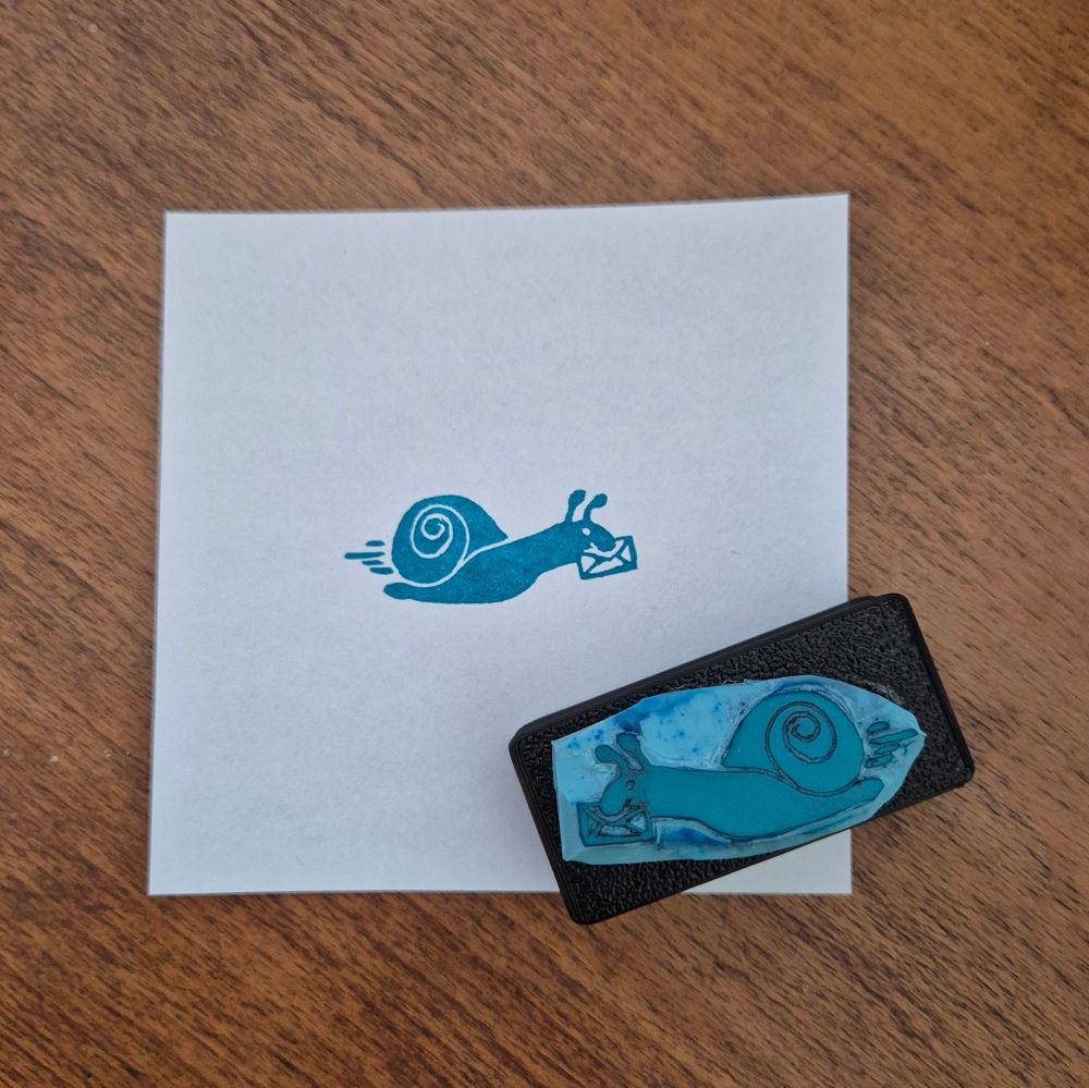 A handcarved rubber stamp and its print in teal showing a snail with a letter in its mouth. 