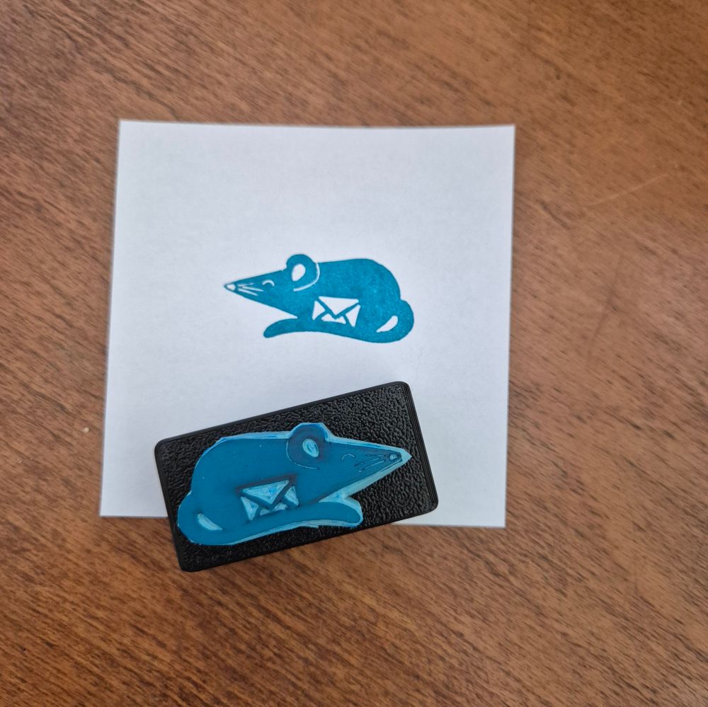 A hand carved rubber stamp and its print in teal of a little mouse with a letter between it and its tail