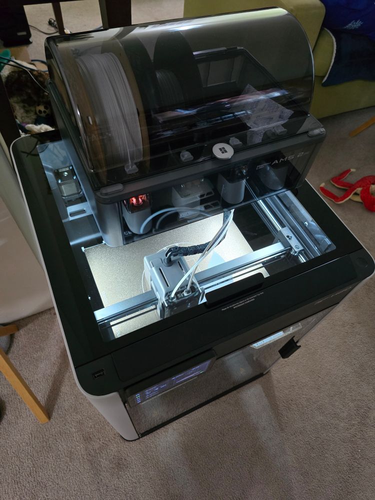 A 3/4ths view of a Bambu Lab H2D 3D printer on a carpeted living room floor.